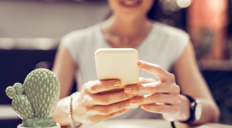 Mulher desfocada ao fundo olhando para o celular que está ofocado à frente da imagem.