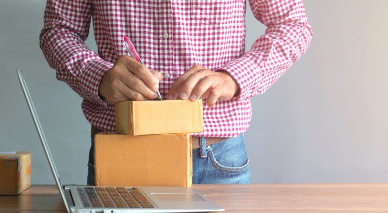 Homem escrevendo em caixas de encomenda ao lado de um notebook aberto