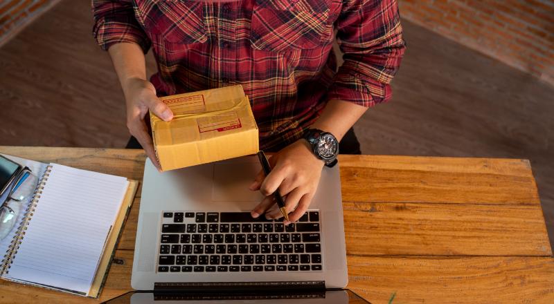 homem com uma caixa de encomenda em uma das mãos e mexendo no notebook