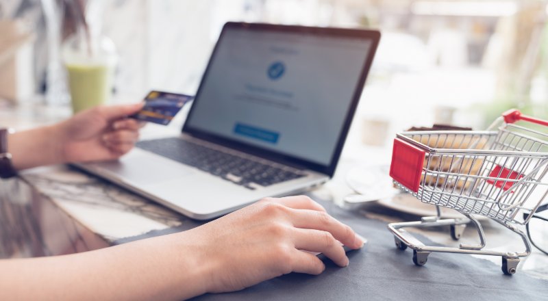 Homem mexendo no notebook ,com o cartão d crédito em uma das mãos e um mini carrinho de compras ao lado 