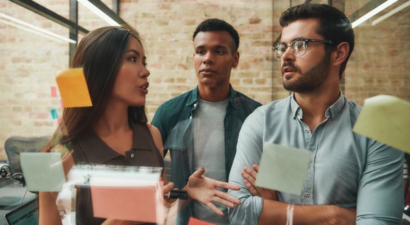 Dois homens e uma mulher debatendo em frente a uma parede transparente com alguns postites grudados.