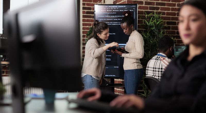 Duas mulheres jovens, mexendo em um tablet com semblante de curiosidade, em um ambiente de escritório com outras pessoas em volta trabalhando em computadores.