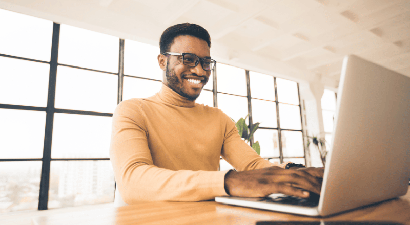 Homem negro, de óculos, mexendo em um notebook e sorrindo. 
