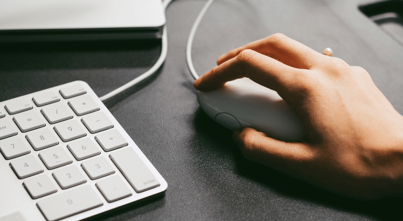 Mão segurando mouse branco e aparece tb um pouco do teclado branco também.