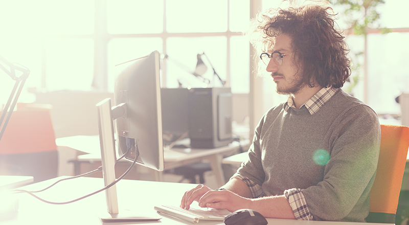 Homem branco, jovem, usando óculos de cabelos e barba castanha, mexendo no computador. Ele está de perfil