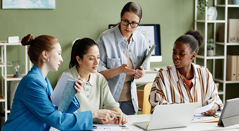 Grupo de mulheres analisando algo em uma tela de notebook que está virada de costas para a foto.