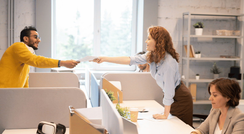 Ambiente de escritório. Homem negro entrega folhas para uma mulher branca, mostrando que uma cultura SEO First pode beneficiar uma empresa.