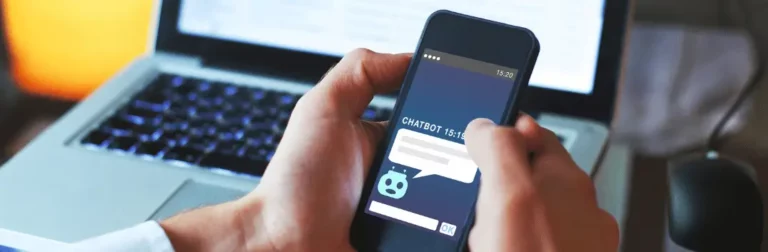 Imagem mostra homem com um celular nas mãos, trocando mensagens com um chatbot e um notebook aberto em segundo plano.