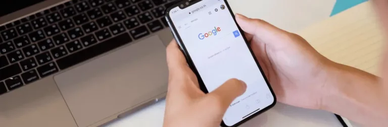 Pessoa com o celular nas mãos, na página inicial do Google. Há um notebook em segundo plano