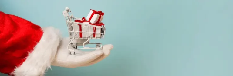 Imagem mostra uma parte do braço do Papai Noel, com um carrinho de compras, contendo presentes, na palma de sua mão.