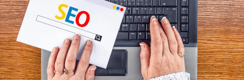 Imagem mostra mãos femininas no teclado de um notebook, segurando um papel escrito SEO com as cores do Google.
