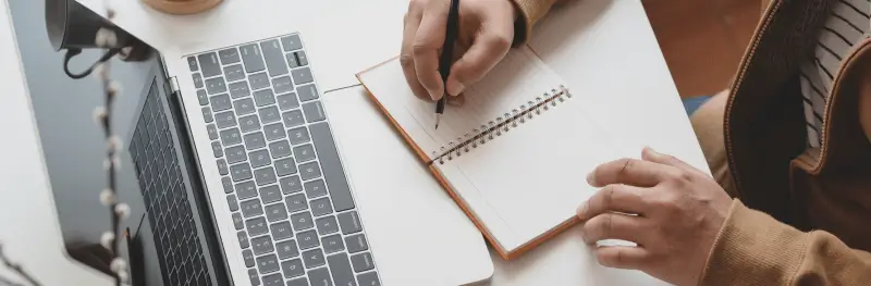 Imagem mostra homem na frente de um notebook aberto, anotando algo em uma agenda