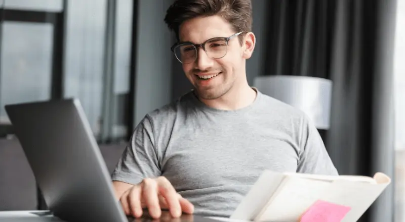 Imagem de um homem sorrindo, olhando para a tela do notebook, digitando e com um caderno na outra mão
