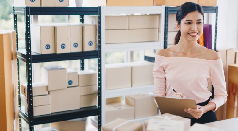 Guia de E-commerce 2022: alavanque sua loja virtual com as dicas Webshare Mulher com uma prancheta em um estoque de produtos embalados
