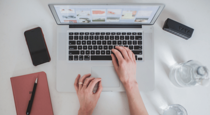 Google Tag Manager: Saiba a importância para seu site e e-commerce Imagem de cima para baixo mostrando mãos de uma pessoa teclando em um notebook. Aparecem também na imagem: um celular uma caneta e um caderninho.