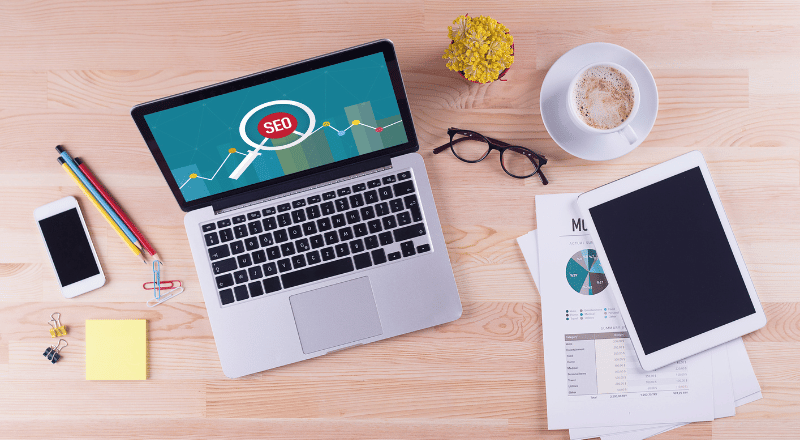 SEO para E-commerce: aprenda a otimizar sua loja virtual Notebook, tblet, café e diversos outros objetos em cima de uma mesa.