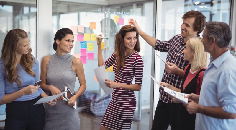 Planejamento Estratégico: Como fazer dicas e exemplos Grupo de funcionários sorrindo e escrevendo em postites grudados em uma parede transparente.