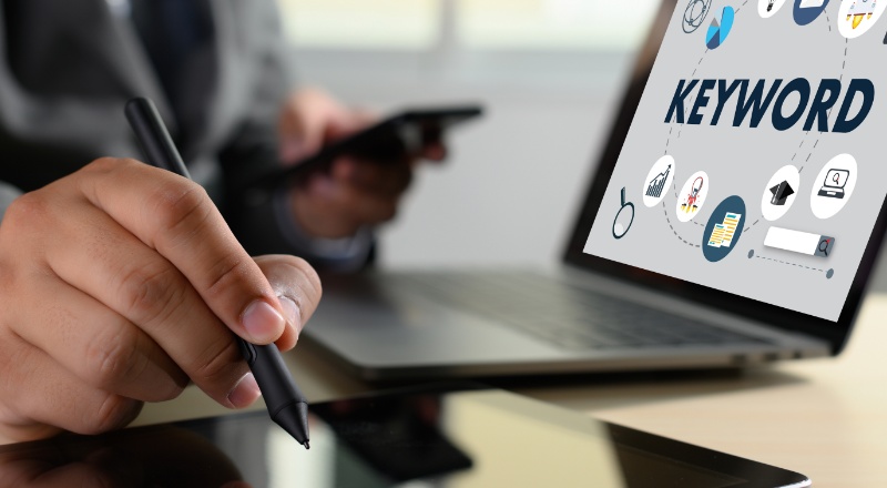 A importância das Branded Keywords para a Estratégia de SEO Homem com um celular em uma mão, mexendo no tablet e o notebook aberto em sua frente, com Keyword escrito na tela
