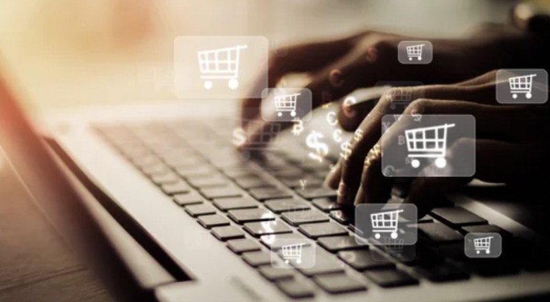 E-commerce no Brasil cresce em 2022 em comparação com o ano passado Duas mãos paertam nas teclas do teclado de um computador. Aparecem ícones de carrinhos de supermercados translúcidos.